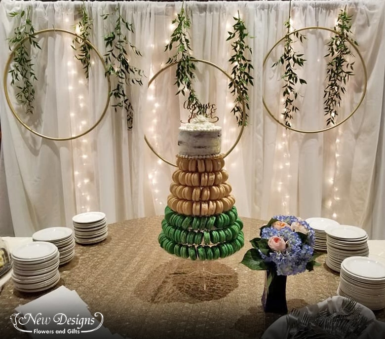 A backdrop sits behind a cake table, white ruffled chiffon with twinkle lights peeking through, 3 hoops decorated with greenery sit at various heights.  A gold sequined table cloth decorates the table, and a throw bouquet of blue hydrangea and peach roses and berries offsets a tall cake stand of Ombre green to white macarons topped with a small white cake.